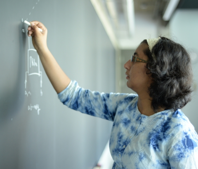 Side profile of a woman wearing a blue tie dye shirt writing on a blackboard