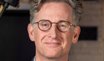 Portrait of a man with greying hair wearing dark rimmed glasses and a suit