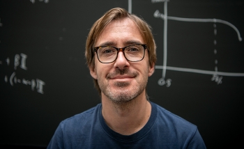 Portrait of a man wearing dark glasses and a blue shirt standing in front of a chalkboard of equations