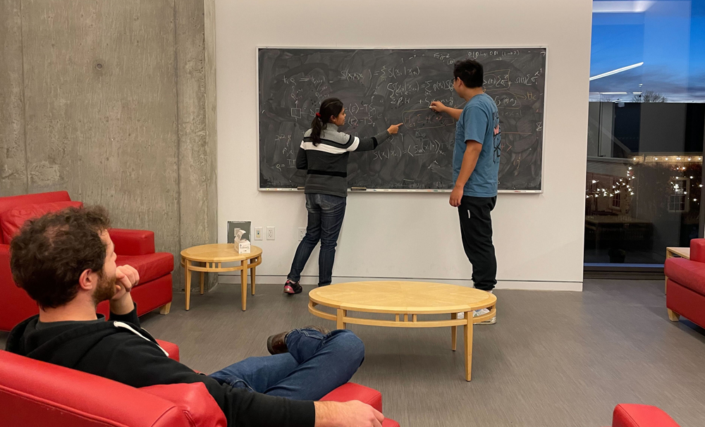 Man and woman working at a blackboard together while another man in a chair watches