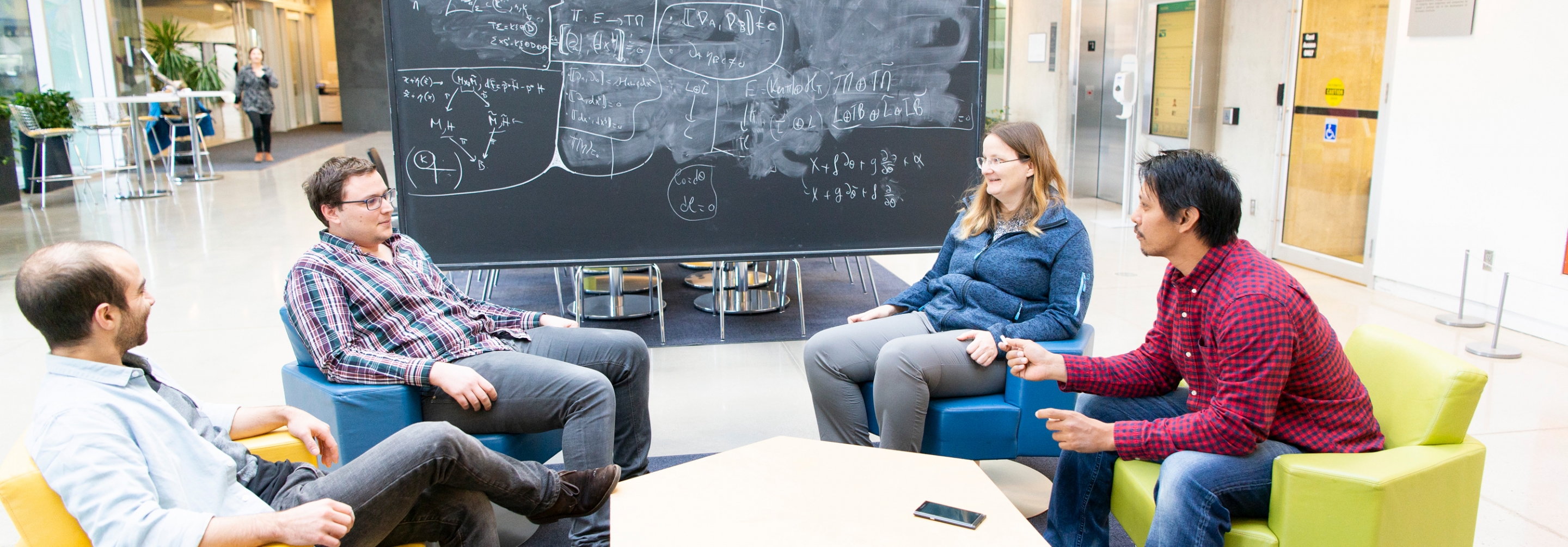 Three men and a woman sitting in a group in front of a chalkboard having a discussion