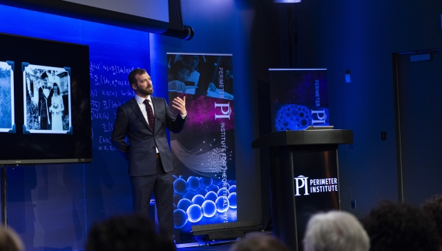 Man wearing a suit and standing in front of an audience giving a lecture